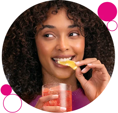 Uśmiechnięta kobieta trzyma w ręce szklankę z gazowanym napojem i gryzie plasterek pomarańczy